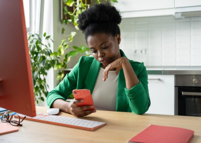 A woman procrastinating on her phone while sitting in front of a computer.